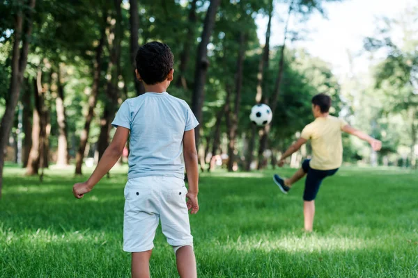 Вид сзади на африканского ребенка, стоящего рядом с мальчиком, играющим в футбол — стоковое фото