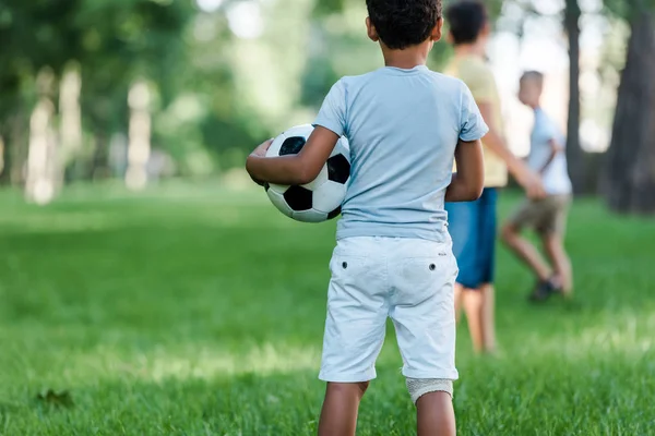 Vista posterior del niño afroamericano de pie con el fútbol cerca de los niños - foto de stock