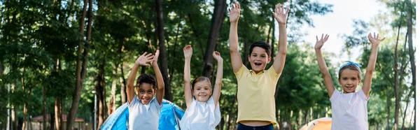 Panoramic shot of happy multicultural kids with hands above heads — Stock Photo