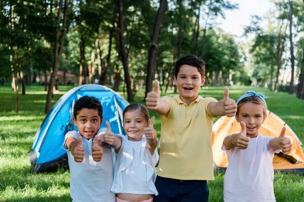 Niños multiculturales felices mostrando pulgares cerca de los campamentos - foto de stock