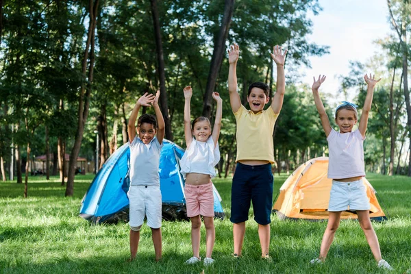 Happy multicultural kids with hands above heads near camps — Stock Photo