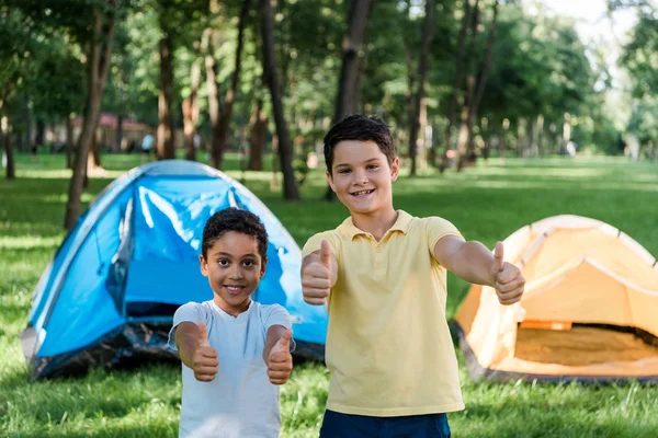 Feliz multicultural chicos sonriendo mientras mostrando pulgares hacia arriba cerca de campamentos - foto de stock