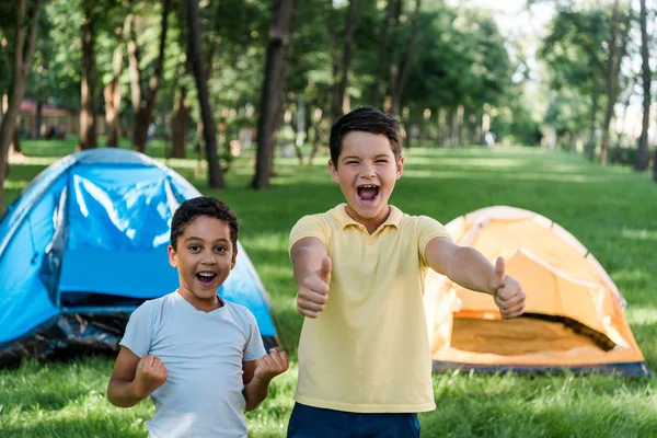 Menino feliz sorrindo ao mostrar polegares perto de amigos e acampamentos afro-americanos — Fotografia de Stock