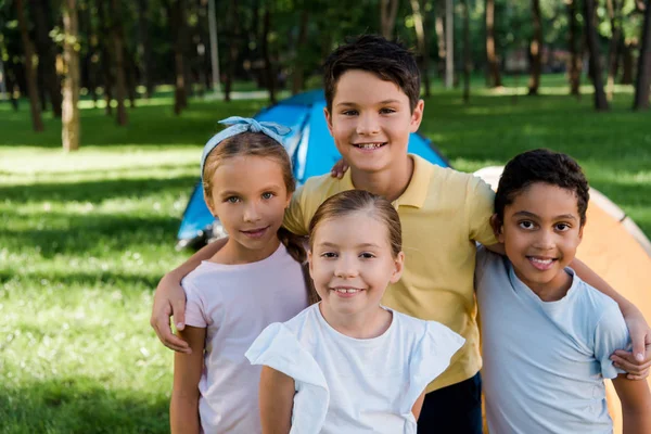 Heureux enfants multiculturels souriant près des camps dans le parc — Photo de stock