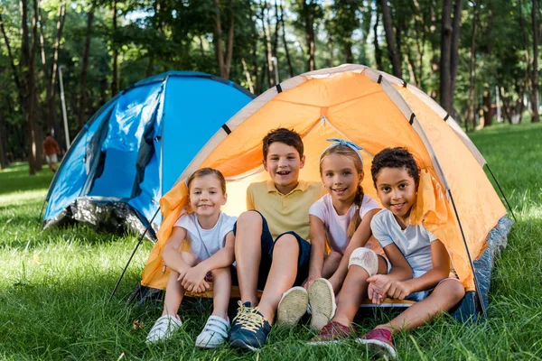 Fröhliche multikulturelle Kinder lächeln, während sie im gelben Lager sitzen — Stockfoto