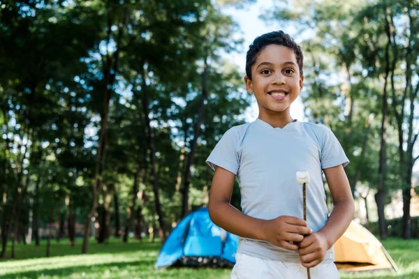 Feliz afroamericano chico sosteniendo palo con dulce malvavisco cerca de campamentos - foto de stock