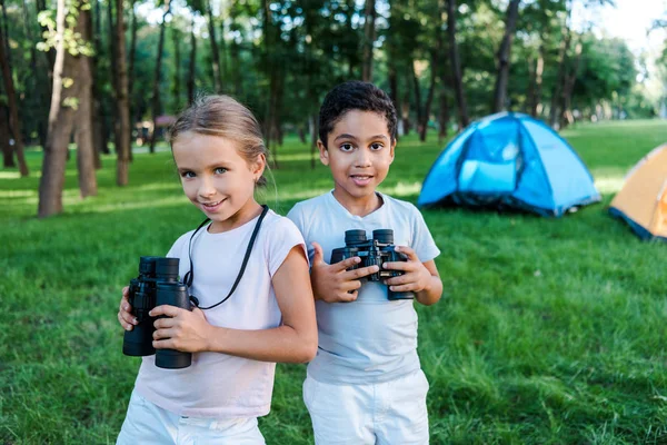 Happy multicultural kids holding binoculars near camp — Stock Photo