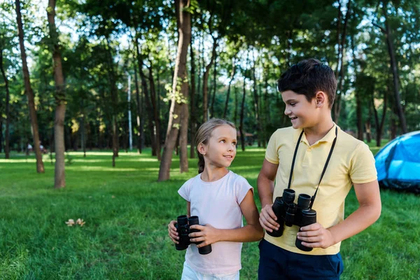 Feliz chico mirando lindo amigo sosteniendo binoculares en parque - foto de stock