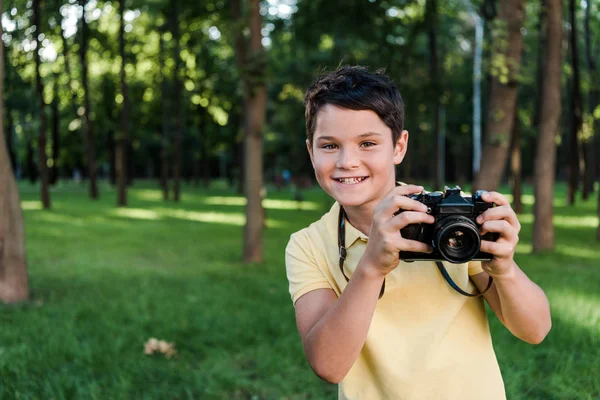 Счастливый мальчик держит цифровой фотоаппарат и улыбается в парке — стоковое фото