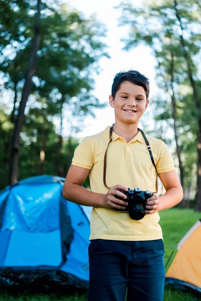Happy boy holding digital camera and smiling near camps in park — Stock Photo
