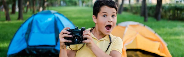 Tiro panorâmico de menino surpreso segurando câmera digital perto de acampamentos no parque — Fotografia de Stock