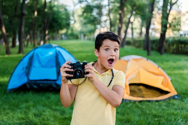 Menino surpreso segurando câmera digital perto de acampamentos no parque — Fotografia de Stock