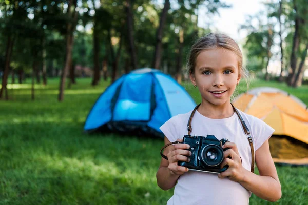 Criança sorrindo segurando câmera digital perto de acampamentos no parque — Fotografia de Stock