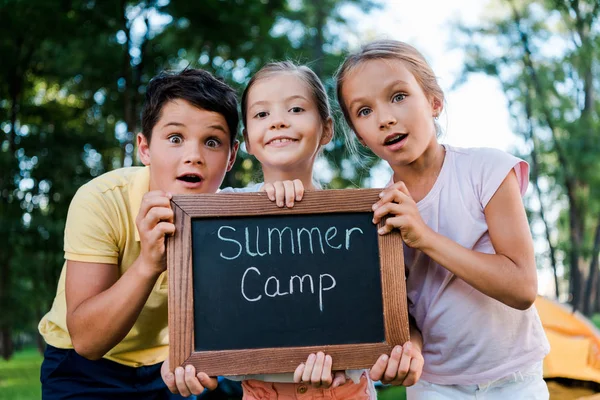 Niños sorprendidos sosteniendo pizarra con letras de campamento de verano - foto de stock