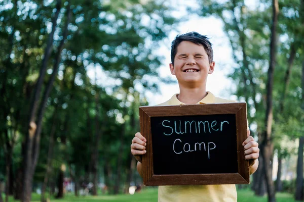 Niño feliz con los ojos cerrados sosteniendo pizarra con las letras del campamento de verano - foto de stock