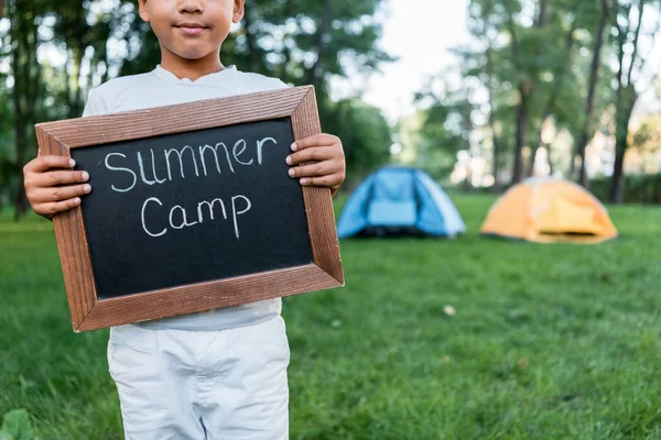 Vista recortada de un niño afroamericano sosteniendo pizarra con letras de campamento de verano - foto de stock