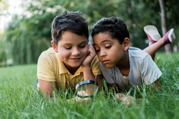 Selective focus of cute multicultural boys lying on grass and looking through magnifier — Stock Photo