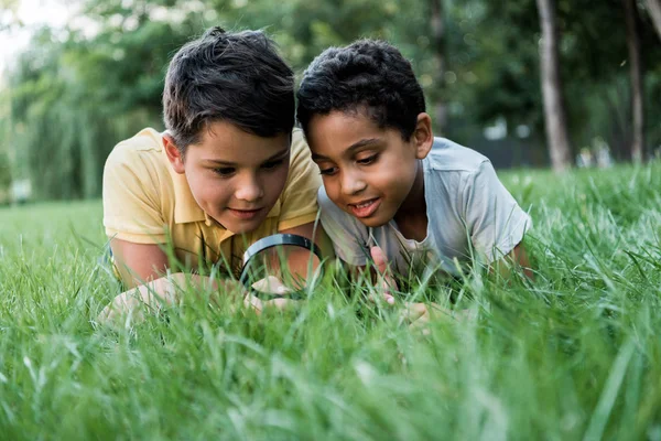 Selective focus of multicultural boys lying on grass and looking through magnifier — Stock Photo
