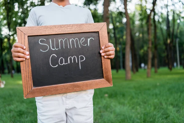 Vue recadrée de gamin afro-américain tenant un tableau craie avec des lettres de camp d'été dans le parc — Photo de stock