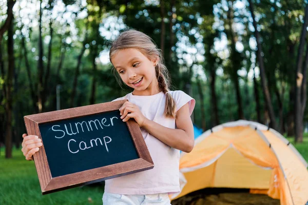 Criança feliz segurando placa de giz com letras de acampamento de verão no parque — Fotografia de Stock