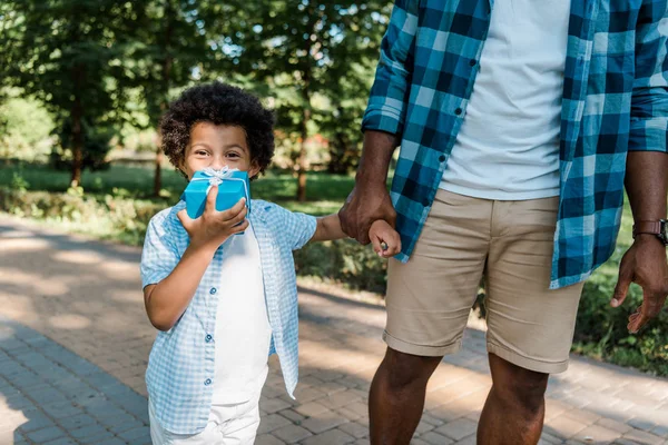 Vista ritagliata dell'uomo afro-americano che si tiene per mano con carino figlio che copre il viso con presente — Foto stock