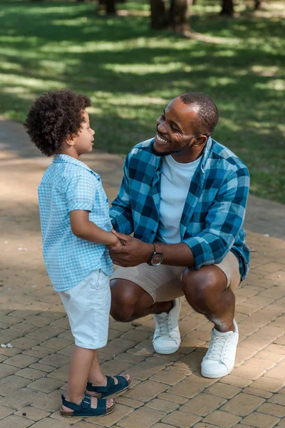 Alegre africano americano hombre cogido de la mano con lindo hijo en parque - foto de stock
