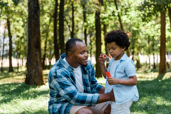 Красивый африканский отец-американец смотрит на милого сына, дующего мыльный пузырь. — стоковое фото