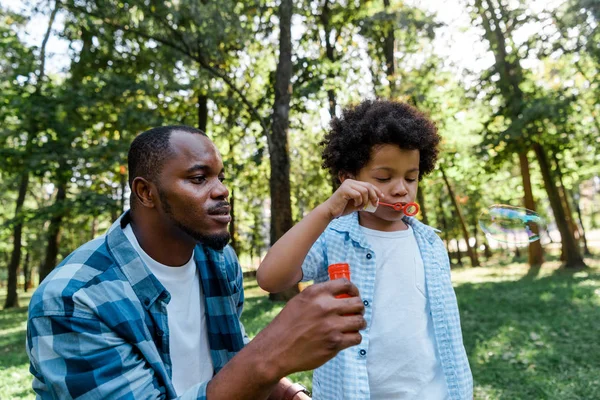 Guapo afroamericano padre mirando adorable hijo soplando jabón burbuja - foto de stock