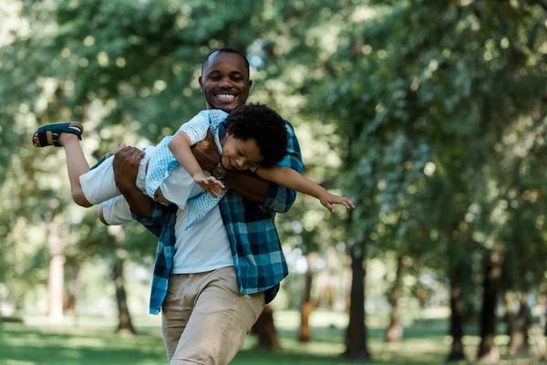 Lächelnder afrikanisch-amerikanischer Vater hält seinen süßen Sohn im grünen Park in den Armen — Stockfoto