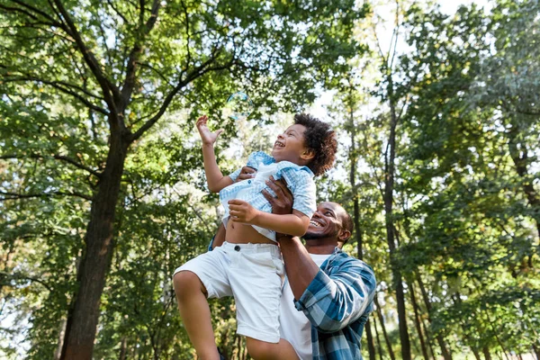 Bajo ángulo vista de feliz afroamericano hombre sosteniendo en brazos lindo hijo en parque - foto de stock