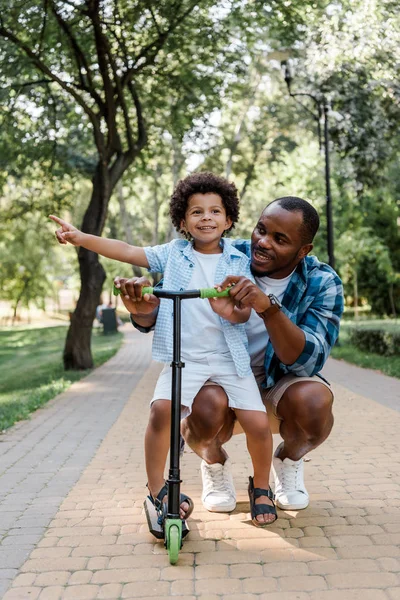 Sorridente padre afroamericano guardando il figlio carino che punta con il dito vicino scooter — Foto stock