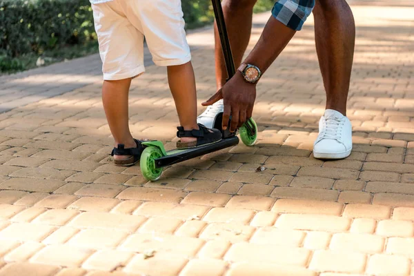 Vista recortada de padre afroamericano tocando scooter cerca de hijo - foto de stock