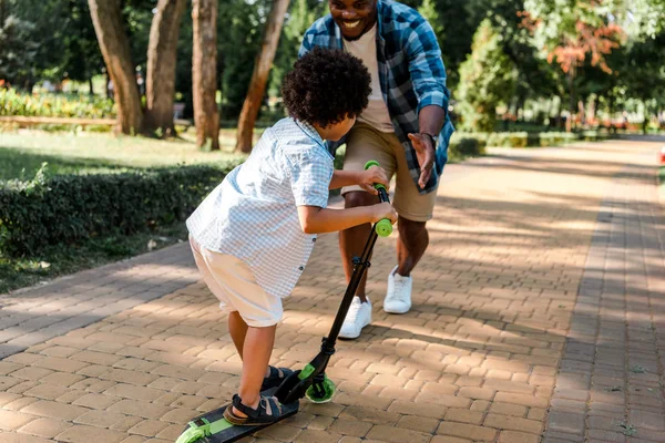 Vue recadrée de heureux père afro-américain gesticulant et courant à son fils sur scooter — Photo de stock