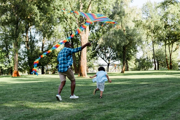 Père afro-américain tenant cerf-volant près du fils bouclé dans le parc — Photo de stock