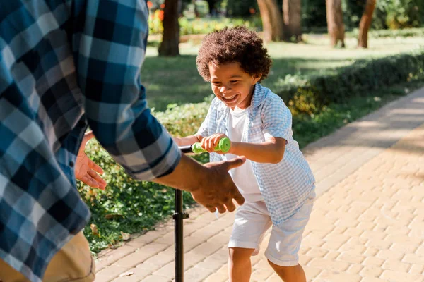 Vista ritagliata del padre gesticolando vicino felice africano americano figlio equitazione scooter — Foto stock