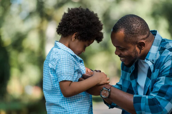 Vue latérale du père afro-américain joyeux tenant la main avec son fils triste — Photo de stock