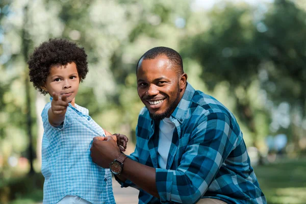 Feliz africano americano hombre mirando cámara cerca lindo hijo apuntando con dedo - foto de stock