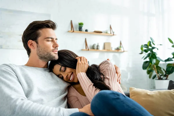 Mujer afroamericana y hombre guapo con los ojos cerrados sonriendo y abrazándose en apartamento - foto de stock