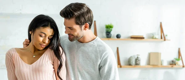 Tiro panorâmico de mulher afro-americana e homem bonito sorrindo e abraçando no apartamento — Fotografia de Stock