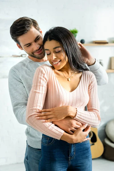 Africano americano mujer y guapo hombre sonriendo y abrazando en apartamento - foto de stock
