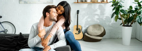 Tiro panorâmico de mulher afro-americana e homem bonito sorrindo e abraçando no apartamento — Fotografia de Stock