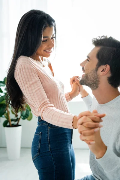 Mujer afroamericana y hombre guapo sonriendo y tomados de la mano en el apartamento - foto de stock