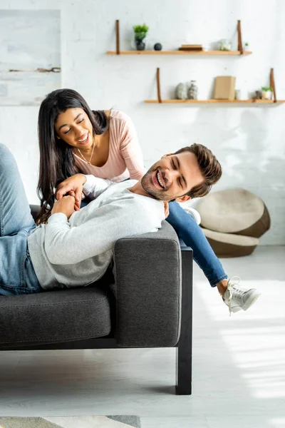 Afro-américaine femme et bel homme souriant et assis sur le canapé dans l'appartement — Photo de stock