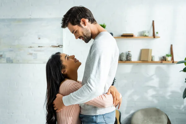 Vista lateral de la mujer afroamericana y el hombre guapo sonriendo y abrazándose en apartamento - foto de stock