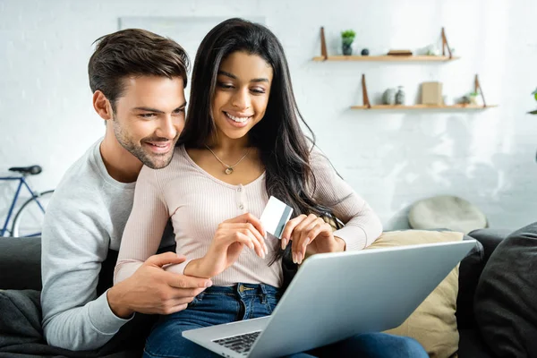 Donna afroamericana sorridente in possesso di carta di credito e guardando il computer portatile con l'uomo — Foto stock