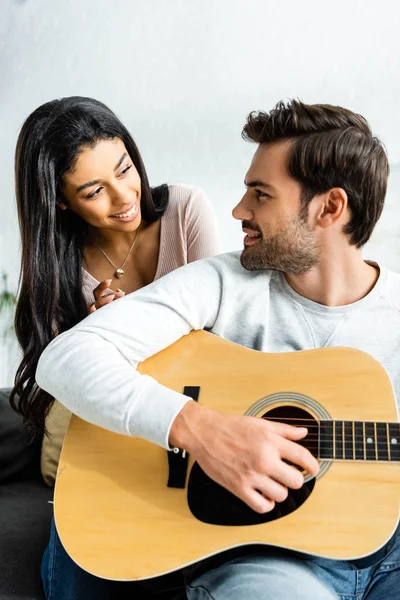 Sonriente mujer afroamericana mirando al hombre con guitarra acústica - foto de stock