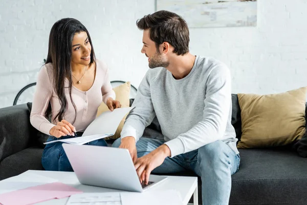 Mujer afroamericana haciendo papeleo y hombre guapo usando portátil en apartamento - foto de stock