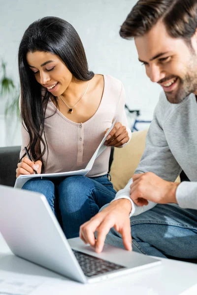 Mujer afroamericana haciendo papeleo y hombre guapo usando portátil en apartamento - foto de stock