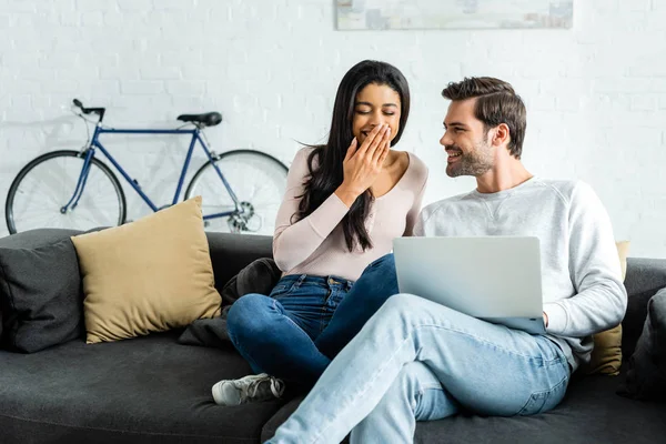 Sonriente afroamericana mujer sentada en sofá y guapo hombre sosteniendo portátil - foto de stock