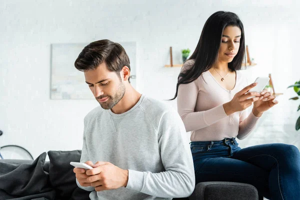 Hombre guapo y mujer afroamericana sentado en el sofá y el uso de teléfonos inteligentes - foto de stock